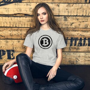 May include: A light gray t-shirt featuring a black Bitcoin symbol. The short-sleeved shirt is worn by a person sitting in front of a wooden background. The person is also wearing black pants and has a red and white helmet.
