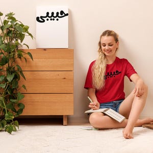 K&ouml;nnte beinhalten: Ein rotes T-Shirt mit schwarzer arabischer Schrift wird von einer Person getragen, die auf einem wei&szlig;en Teppich sitzt. Die gleiche Schrift befindet sich auf einer wei&szlig;en Leinwand auf einer Holzkommode. Links von der Kommode befindet sich eine gr&uuml;ne Pflanze.