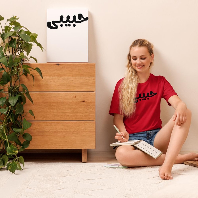 K&ouml;nnte beinhalten: Ein rotes T-Shirt mit schwarzer arabischer Schrift wird von einer Person getragen, die auf einem wei&szlig;en Teppich sitzt. Die gleiche Schrift befindet sich auf einer wei&szlig;en Leinwand auf einer Holzkommode. Links von der Kommode befindet sich eine gr&uuml;ne Pflanze.