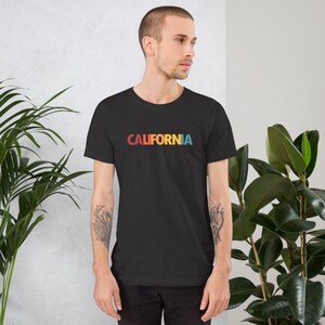 May include: A black t-shirt with the word "CALIFORNIA" printed across the chest in a retro rainbow color scheme. The shirt is short-sleeved and made of a soft material. The person is wearing the shirt.
