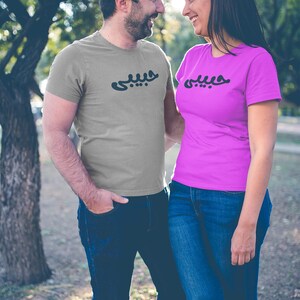 K&ouml;nnte beinhalten: Ein Paar tr&auml;gt passende T-Shirts mit arabischer Schrift. Der Mann tr&auml;gt ein graues Hemd und die Frau ein pinkfarbenes Hemd. Beide Hemden haben die gleiche schwarze Schrift. Sie stehen drau&szlig;en vor einem Baum.