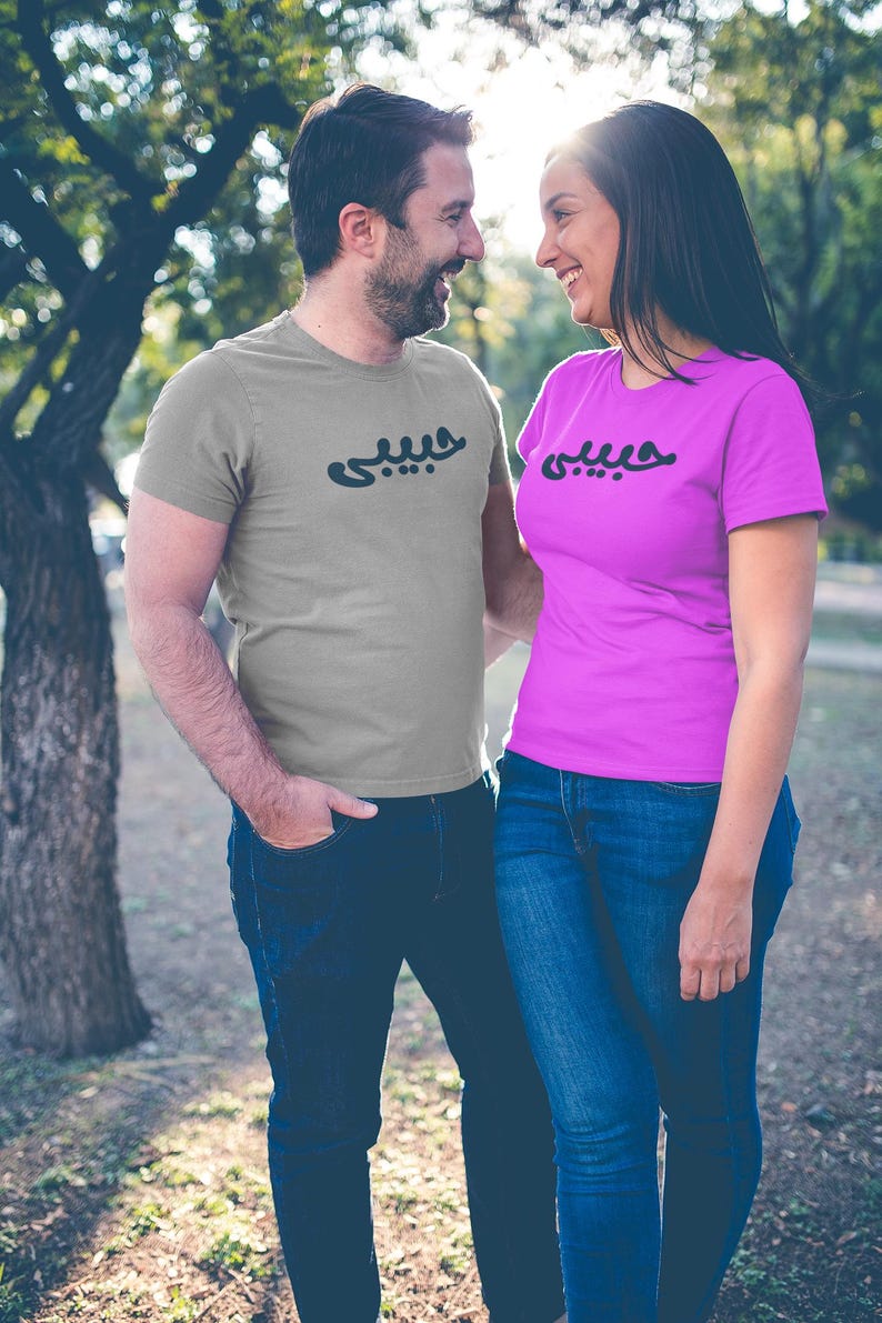 K&ouml;nnte beinhalten: Ein Paar tr&auml;gt passende T-Shirts mit arabischer Schrift. Der Mann tr&auml;gt ein graues Hemd und die Frau ein pinkfarbenes Hemd. Beide Hemden haben die gleiche schwarze Schrift. Sie stehen drau&szlig;en vor einem Baum.