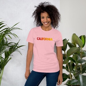 Op de afbeelding: Een lichtroze t-shirt met het woord "CALIFORNIA" in een gradi&euml;nt van rood naar geel. Het shirt met korte mouwen is gemaakt van een zacht materiaal.