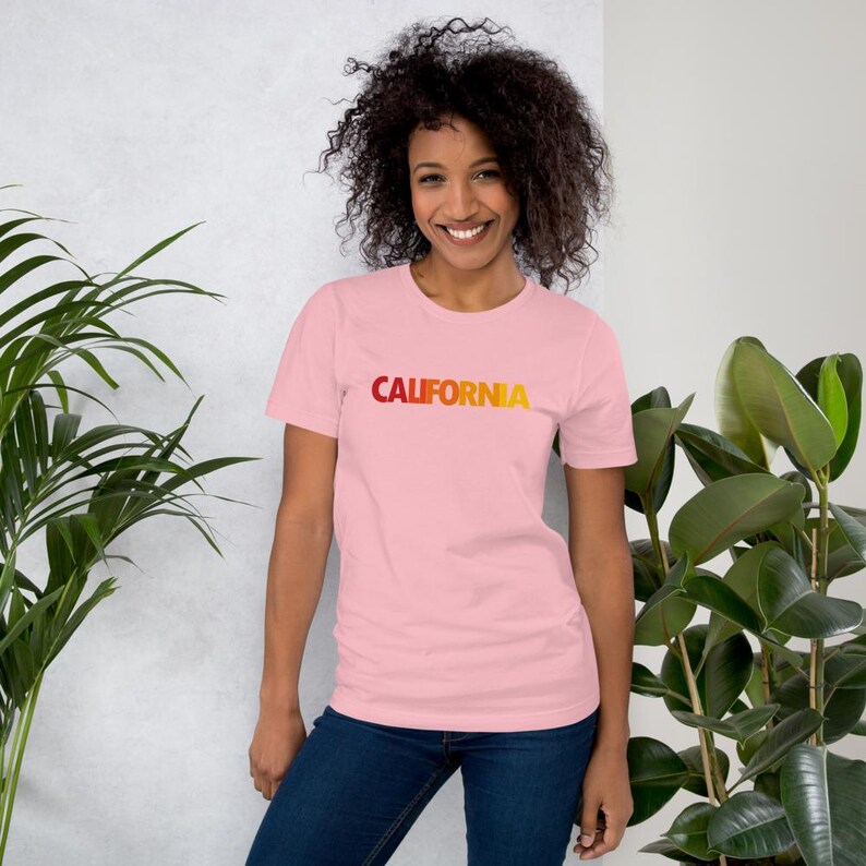 Op de afbeelding: Een lichtroze t-shirt met het woord "CALIFORNIA" in een gradi&euml;nt van rood naar geel. Het shirt met korte mouwen is gemaakt van een zacht materiaal.