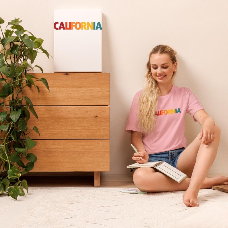 May include: A pink t-shirt with the word "CALIFORNIA" in rainbow colors. A white canvas with the same text is on a wooden dresser. A person is sitting on the floor wearing the t-shirt and writing in a notebook.