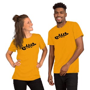 K&ouml;nnte beinhalten: Senfgelbe T-Shirts mit schwarzer arabischer Schrift. Die T-Shirts sind kurz&auml;rmelig und werden von zwei Personen getragen. Der Text auf den Shirts scheint derselbe zu sein. Die Personen l&auml;cheln und stehen vor einem wei&szlig;en Hintergrund.