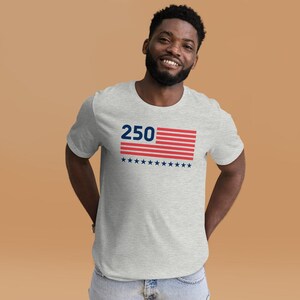 K&ouml;nnte beinhalten: Ein hellgraues T-Shirt mit der Zahl "250" &uuml;ber einem rot-wei&szlig; gestreiften Design und blauen Sternen. Das Shirt ist ein kurz&auml;rmeliges Rundhals-T-Shirt.