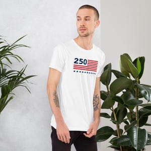 K&ouml;nnte beinhalten: Wei&szlig;es Kurzarm-T-Shirt mit einem grafischen Design. Das Design zeigt die Zahl "250" in Marineblau, &uuml;ber einem rot-wei&szlig; gestreiften Rechteck mit marineblauen Sternen. Das T-Shirt ist aus einem weichen Material.