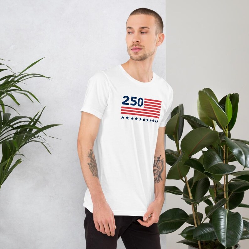K&ouml;nnte beinhalten: Wei&szlig;es Kurzarm-T-Shirt mit einem grafischen Design. Das Design zeigt die Zahl "250" in Marineblau, &uuml;ber einem rot-wei&szlig; gestreiften Rechteck mit marineblauen Sternen. Das T-Shirt ist aus einem weichen Material.