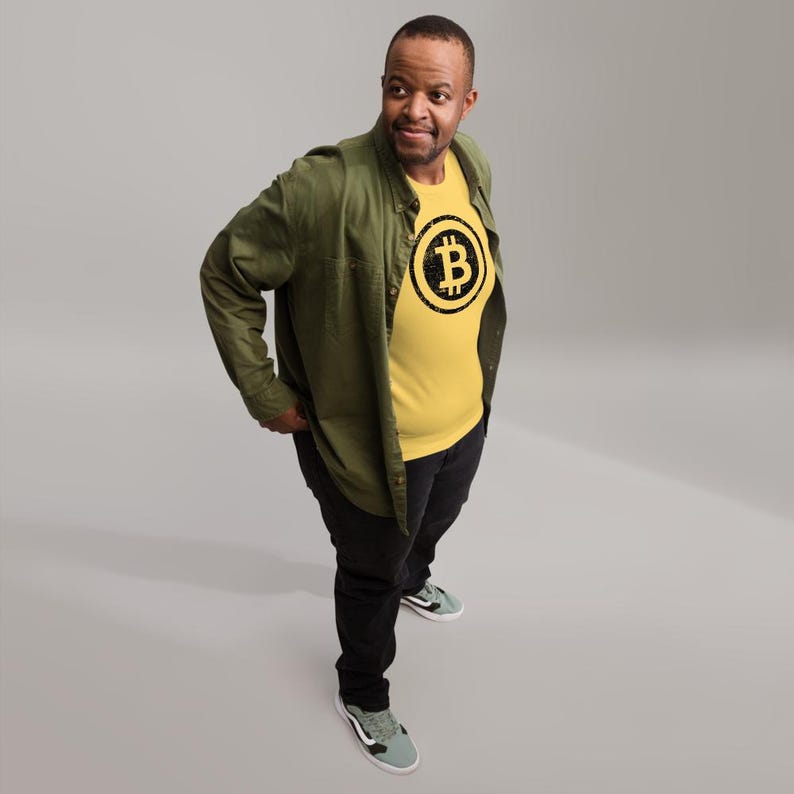 May include: A person wearing a yellow t-shirt with a black Bitcoin symbol graphic. The person is also wearing a green button-down shirt, black pants, and gray sneakers. The background is a solid gray.