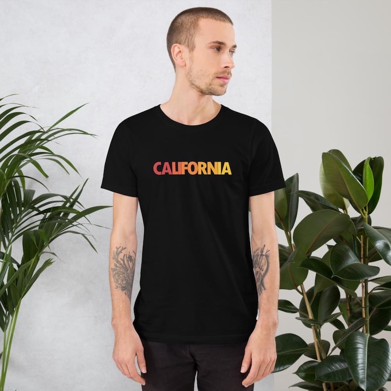 Op de afbeelding: Zwart t-shirt met het woord "CALIFORNIA" in een verloop van rood, oranje en geel. Het shirt met korte mouwen heeft een ronde hals. De tekst staat in het midden op de borst.