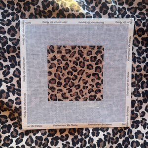 May include: Square needlepoint canvas featuring a leopard print design in shades of brown and tan. The canvas is surrounded by a white border with the text "NEEDLEPOINT BY MEGAN" printed repeatedly. The canvas is placed on a leopard print fabric.