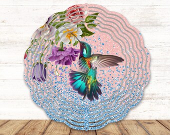 Puede incluir: Un molinillo de viento circular de metal con un colibrí vibrante en vuelo rodeado de flores coloridas. El fondo muestra un efecto de purpurina rosa texturizada que se transforma en un patrón de purpurina azul en el borde inferior.