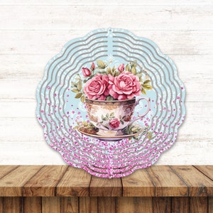 May include: A blue and white wind spinner with a teacup and pink roses. The teacup has a floral pattern and is surrounded by pink glitter.