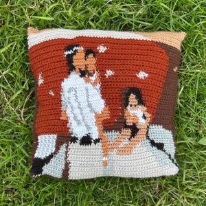 May include: A square, crocheted cushion depicting a family scene. The cushion has a rust-coloured background with a couple and a child depicted in white, tan, and black yarn. The border is a light grey and tan.