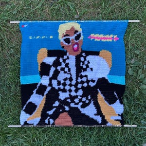 May include: A handmade crochet wall hanging depicting a person in sunglasses and a black and white checkered outfit. The background is blue with pink and yellow text. The artwork is held by two wooden dowels. The piece is made with yarn.