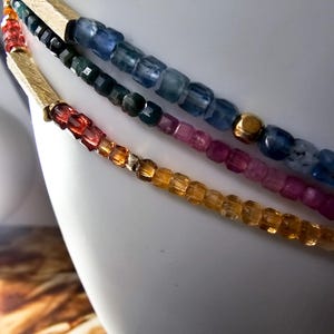 Puede incluir: Primer plano de tres collares de cuentas con detalles dorados. Las cuentas son en tonos azul, verde, rosa, naranja y amarillo. Los collares se muestran sobre una superficie blanca.