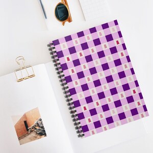 Op de afbeelding: Een spiraalgebonden notitieboek met een paars en roze ruitpatroon, versierd met kleine gummybeer-illustraties. Het notitieboek is open, met lege pagina's en een gouden paperclip. Zonnebril en pen zijn ook zichtbaar.