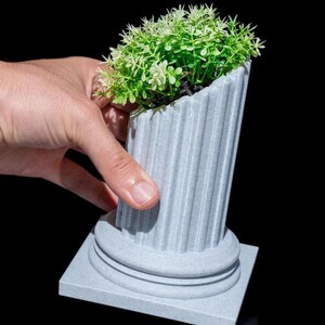 Peut inclure: Un pot de fleurs gris en forme de colonne cannelée contenant une petite plante verte avec des accents blancs. Le pot a une base carrée et est tenu par une main sur fond noir. La plante est d'un vert vif.