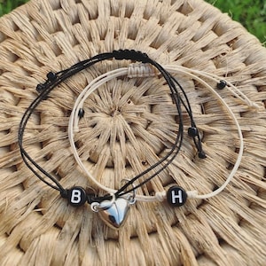 May include: Two friendship bracelets made with black and white string, a silver heart charm, and the letters "B" and "H" on the charms.