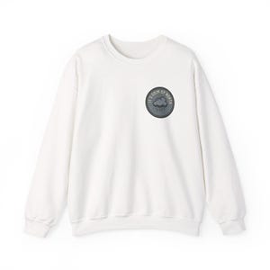 May include: A white long-sleeve sweatshirt featuring a circular patch on the upper left chest. The patch is dark gray with a stitched border and displays a graphic of a rain cloud with the text IT'S GRIM UP NORTH printed in a circular arc.