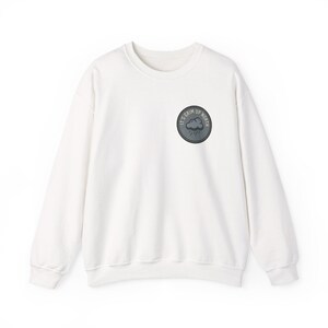 May include: A white long-sleeve sweatshirt featuring a circular patch on the upper left chest. The patch is dark gray with a stitched border and displays a graphic of a rain cloud with the text IT'S GRIM UP NORTH printed in a circular arc.