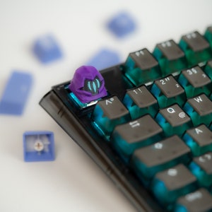 May include: A purple and blue keyboard keycap with a helmet design. The keycap is on a black mechanical keyboard with blue and green keycaps.