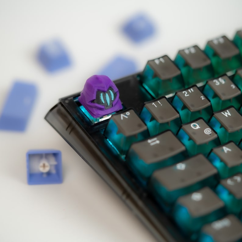 May include: A purple and blue keyboard keycap with a helmet design. The keycap is on a black mechanical keyboard with blue and green keycaps.
