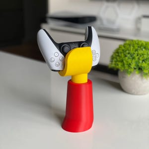 Könnte beinhalten: Ein weißer und schwarzer Videospiel-Controller wird von einem gelben und roten Ständer aufrecht gehalten. Der Ständer hat eine abgerundete Basis und eine gelbe Oberseite, die den Controller hält.