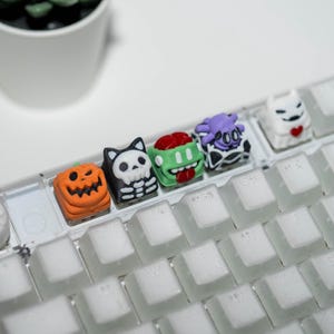 Könnte beinhalten: Eine Sammlung farbenfroher, neuartiger Tastenkappen mit Halloween-Motiven. Die Tastenkappen zeigen einen Kürbis, eine Totenkopfkatze, einen Zombie, eine Kuh und einen Geist. Die Tastenkappen befinden sich auf einer weißen Tastatur.