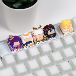 Op de afbeelding: Vijf kleurrijke toetsen met anime-personages met verschillende kapsels en outfits. De toetsen zijn geplaatst op een wit mechanisch toetsenbord.