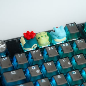 May include: A keyboard with blue and black keys featuring three small, colorful, 3D printed keycaps shaped like cartoon characters. The keycaps are labeled with the numbers 1, 2, 3, 4, and 5.