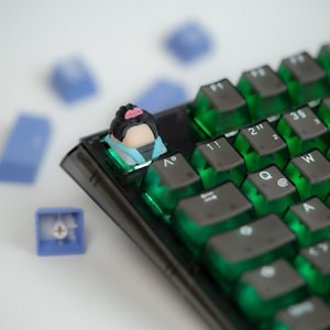 May include: A black mechanical keyboard with green keycaps and a custom keycap featuring a cartoon character with pink hair and a blue and white outfit.