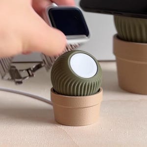 Könnte beinhalten: Ein grüner und brauner Keramik-Ladeständer für eine Apple Watch. Der Ständer ist in Form einer kleinen Topfpflanze gestaltet. Die Uhr wird auf dem Ständer geladen.