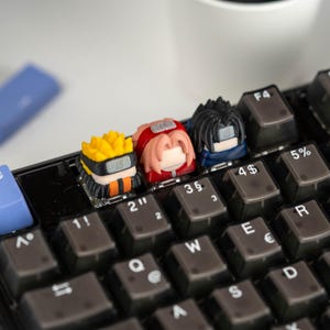 May include: A black mechanical keyboard with custom anime-themed keycaps. The keycaps feature characters with yellow, red, and black hair. The keyboard has grey keys with white lettering and symbols. The keyboard is a compact design.