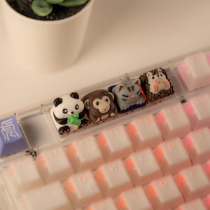 Może przedstawiać: Biała klawiatura z rzędem czterech klawiszy z motywami zwierząt. Klawisze to panda, małpa, kot i chomik. Klawiatura ma różowe podświetlenie.