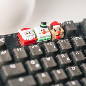 Könnte beinhalten: Drei Weihnachts-Tastenkappen für eine Tastatur. Die Tastenkappen sind aus Kunststoff und zeigen einen Schneemann, den Weihnachtsmann und ein Rentier. Die Tastenkappen befinden sich auf einer schwarzen Tastatur mit weißen Beschriftungen.