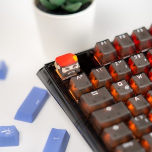 May include: A close-up of a black mechanical keyboard with a custom keycap featuring a cartoon character with a red hat and a beard. The keycap is on the "Esc" key. The keyboard has orange backlighting.