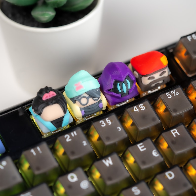 May include: Four colorful keycaps with different designs, including a purple robot, a blue and white character, a yellow and blue character, and a red character with a mustache, are placed on a black keyboard with yellow keys. The keys are labeled with numbers and symbols, including 11, 2", 3&sect;, 4$, 5%, 2, 3, W, S, E, &euro;, D, and R.