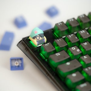 May include: A green and yellow character keycap with glasses sits on a black keyboard with green keys. The keys have symbols like "!" and "@" on them.