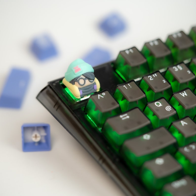 May include: A green and yellow character keycap with glasses sits on a black keyboard with green keys. The keys have symbols like "!" and "@" on them.