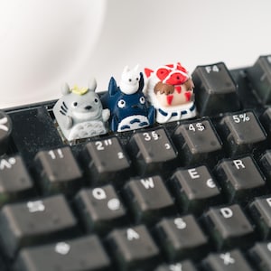 Può includere: Quattro piccole figurine stampate in 3D di personaggi del film d'animazione 'Il mio vicino Totoro' sono posizionate su una tastiera nera. Le figurine sono posizionate sui tasti contrassegnati '2', '3', '4' e '5'.