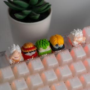 Op de afbeelding: Vijf kleurrijke keycaps met anime-personages op een wit toetsenbord met rode achtergrondverlichting. De keycaps zijn gemaakt van hars en hebben gedetailleerde ontwerpen.
