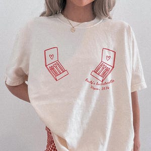 May include: An ivory short-sleeved t-shirt featuring a red line-art graphic of two open matchboxes with heart symbols. The text "Emily's bachelorette Vegas, 2026" is printed in red cursive script below the right matchbox graphic on the front.