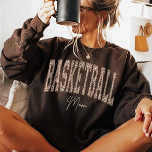 May include: A dark chocolate brown crewneck sweatshirt featuring the word BASKETBALL in a leopard print pattern across the chest, with the word Mom written in a cursive script below it and the name Olivia on the sleeve near the wrist.