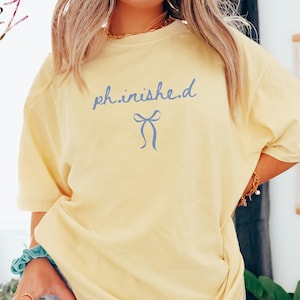 May include: A light yellow t-shirt with the text "ph.inished" in light blue script and a blue bow graphic. The shirt is a relaxed fit and has short sleeves. The word "Banana" is in a white box in the bottom right corner.