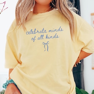 May include: A pale yellow t-shirt featuring the blue text "celebrate minds of all kinds" and a blue bow graphic. The shirt has a loose fit and short sleeves, suitable for casual wear.