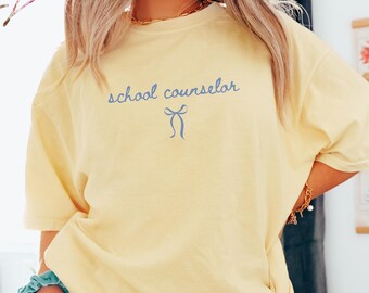 Könnte beinhalten: Hellgelbes T-Shirt mit dem blauen Schriftzug "school counselor" und einer blauen Schleifengrafik. Das Shirt hat eine lockere Passform. Das Wort "Banana" befindet sich in einer weißen Box unten rechts.