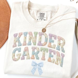 May include: Cream-coloured t-shirt featuring the word "KINDERGARTEN" in pastel patchwork lettering. A light blue gingham bow is below the text. A gold necklace with a pendant is draped across the shirt. The shirt is folded.