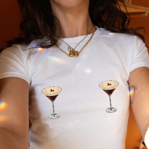 May include: White crew neck t-shirt with two cocktail glass designs. The glasses are filled with a dark liquid, topped with a light foam and a garnish. The shirt is a casual style.